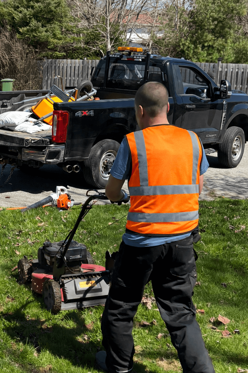 Landscaper in orange safety vest mows green lawn with red mower near black pickup truck.