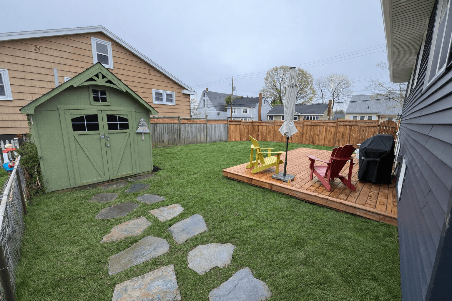 Backyard with green shed, stone path, and wooden deck featuring red and yellow Adirondack chairs.